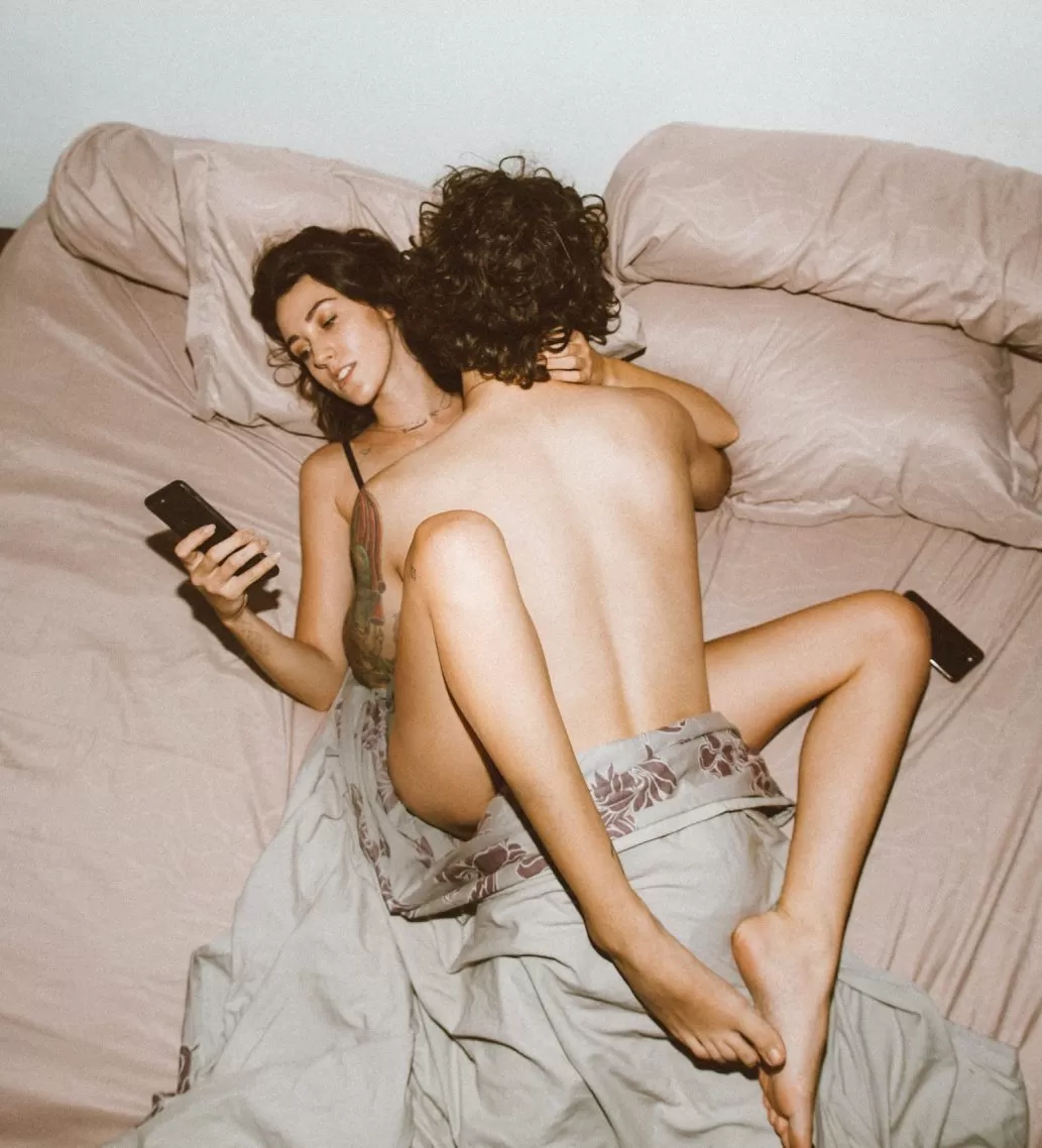 Men on top of a woman in bed while she is on her phone